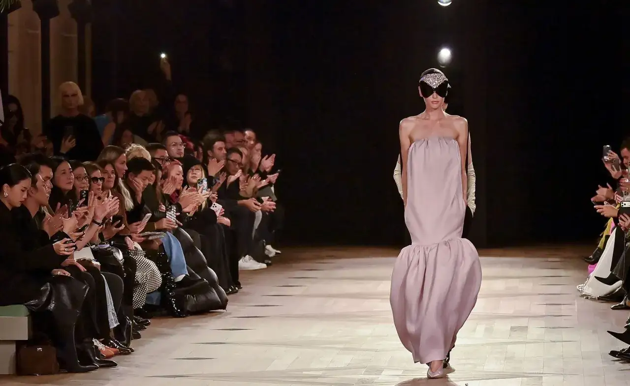 Sfilata Balenciaga: un modello indossa un abito rosa pallido con un'audace maschera scintillante, mentre il pubblico applaude.