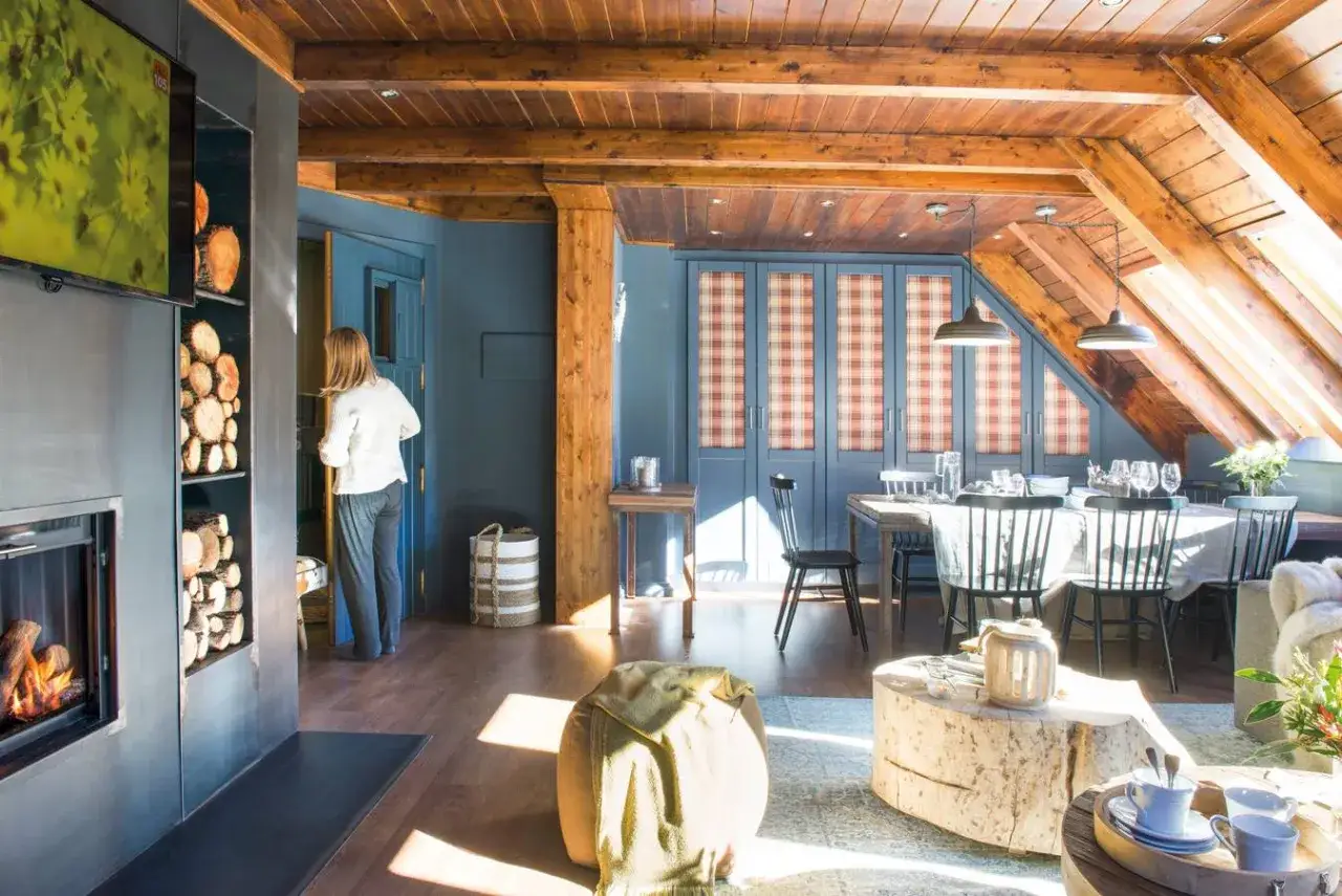 Interior acogedor de una buhardilla con vigas de madera, chimenea, zona de comedor y una mujer de pie.