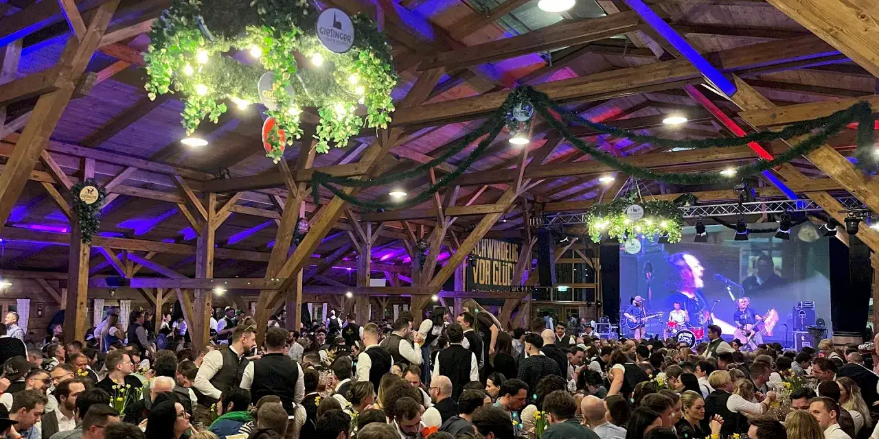 Festzelt-Stimmung beim Nockherberg 2026: Viele Menschen feiern unter Holzbalken, Lichterketten und Girlanden. Eine Band spielt auf der Bühne.