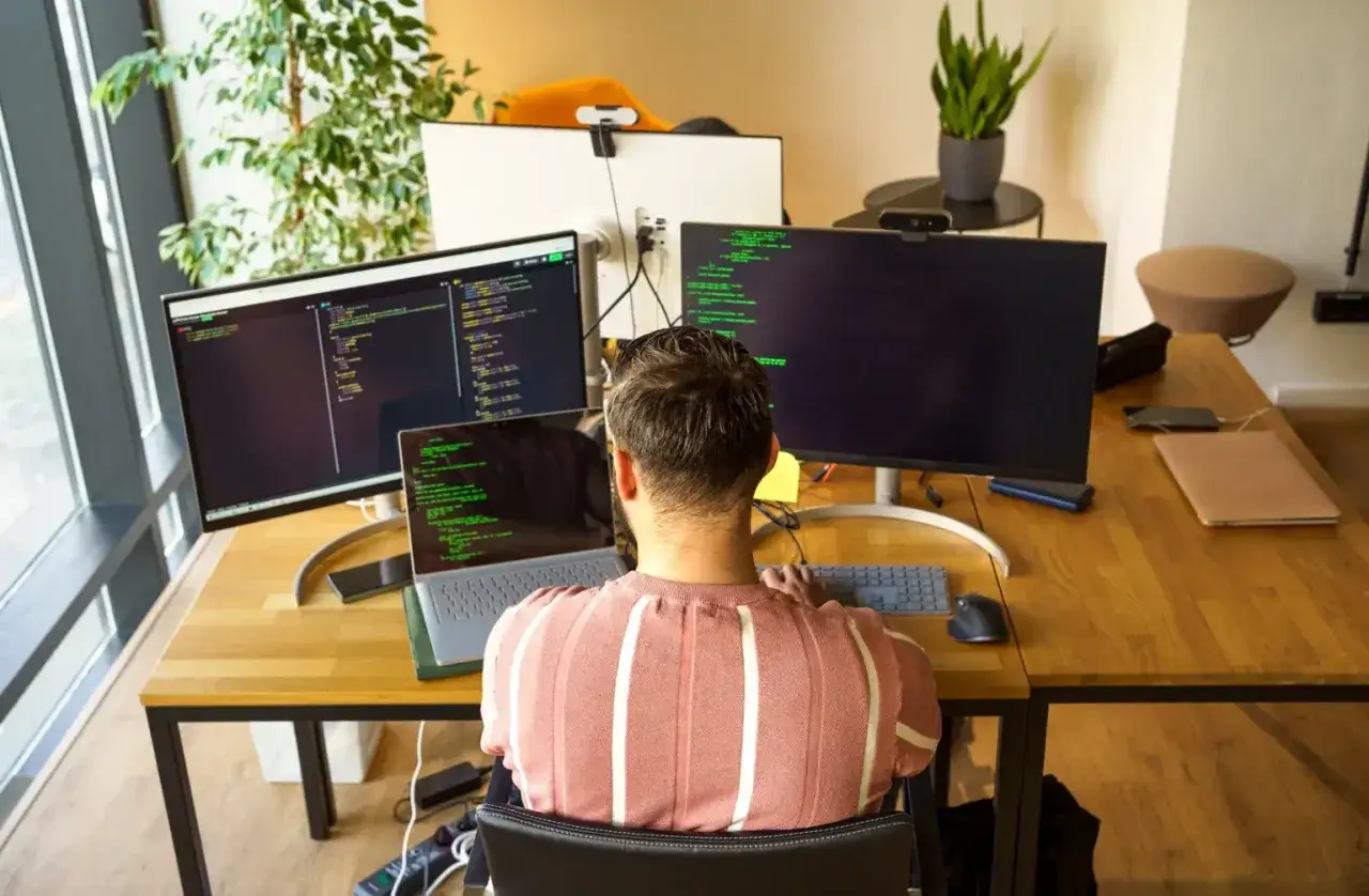 Man kodar vid tre skärmar, en laptop och en tangentbord. Han lär sig nya saker genom e-learning.