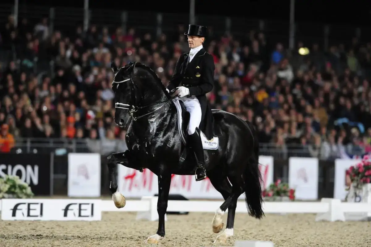 Le célèbre cheval Totilas, noir et brillant, exécute un piaffer majestueux sous son cavalier en tenue de dressage devant une foule attentive.