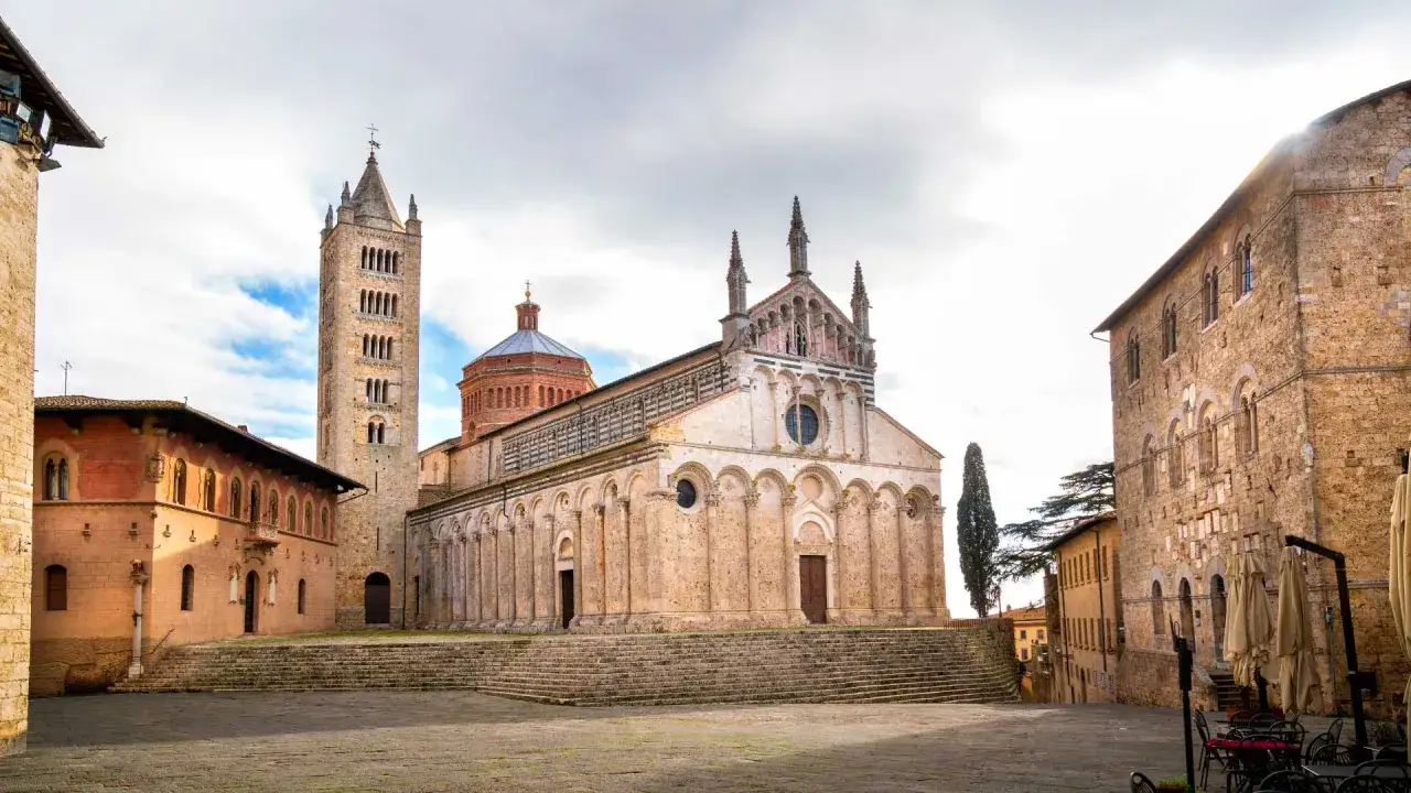 Piazza del Duomo a Massa Marittima: cosa vedere. Maestoso Duomo romanico, torre campanaria e palazzi storici in pietra.