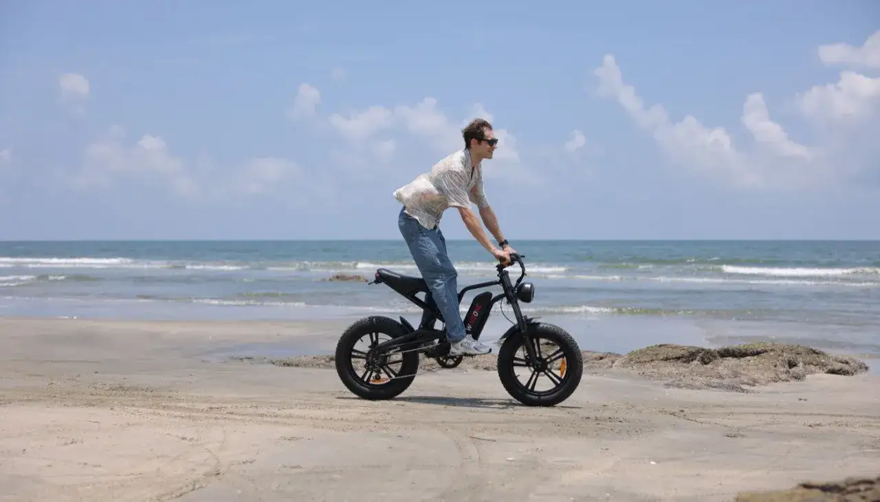 Mężczyzna w okularach przeciwsłonecznych i kwiecistej koszuli jedzie rowerem elektrycznym bez wspomagania po piaszczystej plaży.