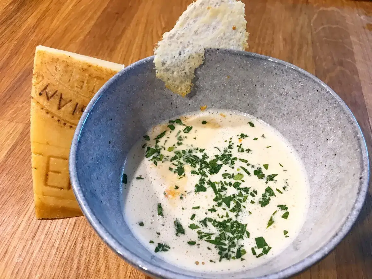 Cremige Suppe mit Kräutern, garniert mit einem knusprigen Parmesan-Chip. Daneben ein Stück Parmesan, dessen Rinde essbar ist.