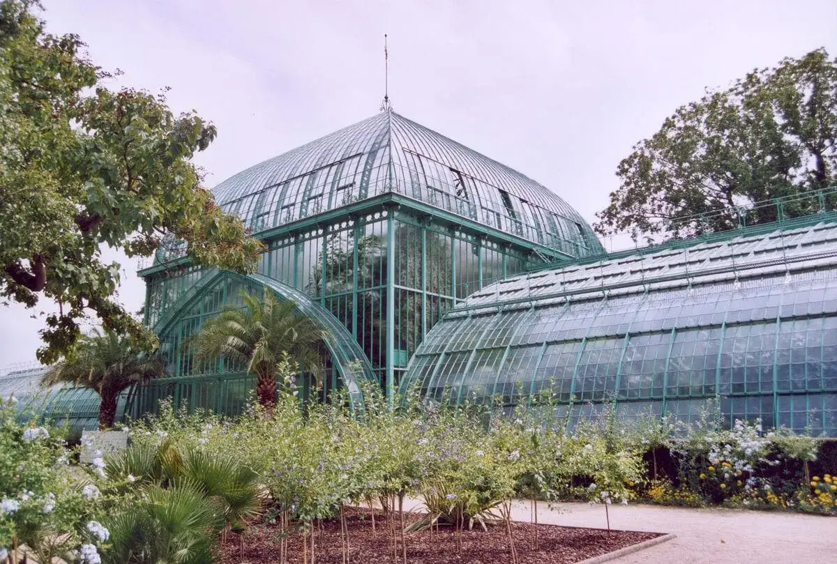 Un impresionante jard&iacute;n de plantas con una gran estructura de cristal verde, palmeras y flores exuberantes.