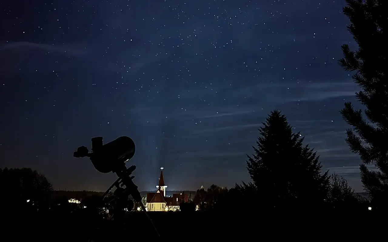 Teleskop na tle nocnego nieba pełnego gwiazd i chmur. W oddali widać oświetlony kościół. Idealne warunki do astronomia studia.