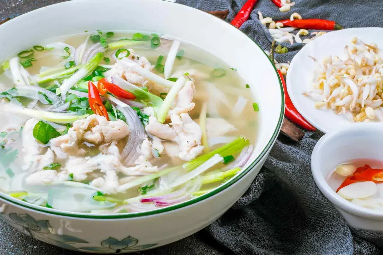 Eine Schüssel mit Canh Chua, einer vietnamesischen Hühnersuppe, garniert mit Frühlingszwiebeln und Chili. Daneben liegen Sprossen und eine kleine Schale mit Knoblauch und Chili.