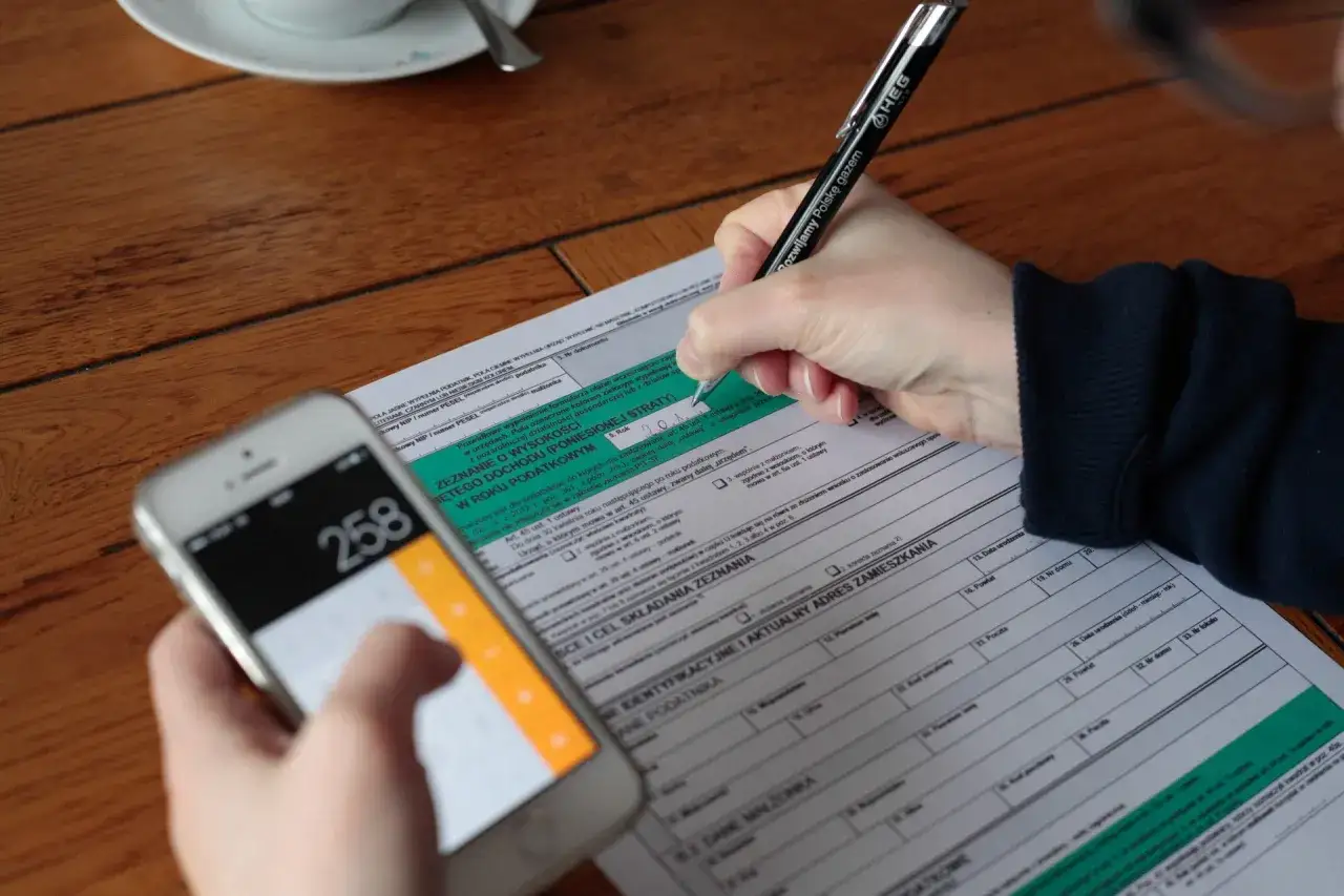 Ręka wypełnia PIT, a obok leży smartfon z kalkulatorem. Czas rozliczyć podatek belki i inne dochody.