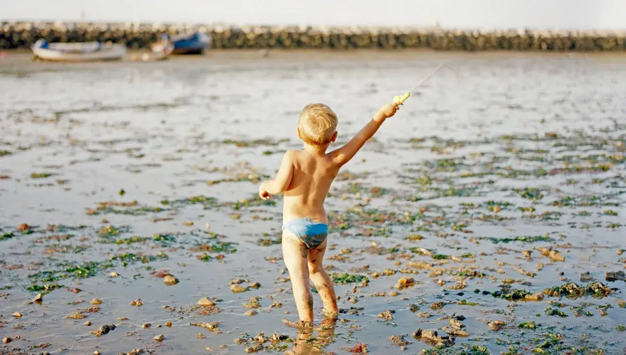 Bambino con canna da pesca in mano, in piedi nell'acqua bassa, con alghe e rocce. Un'immagine che evoca la spensieratezza dell'infanzia, come quelle di **Laura Pannack**.