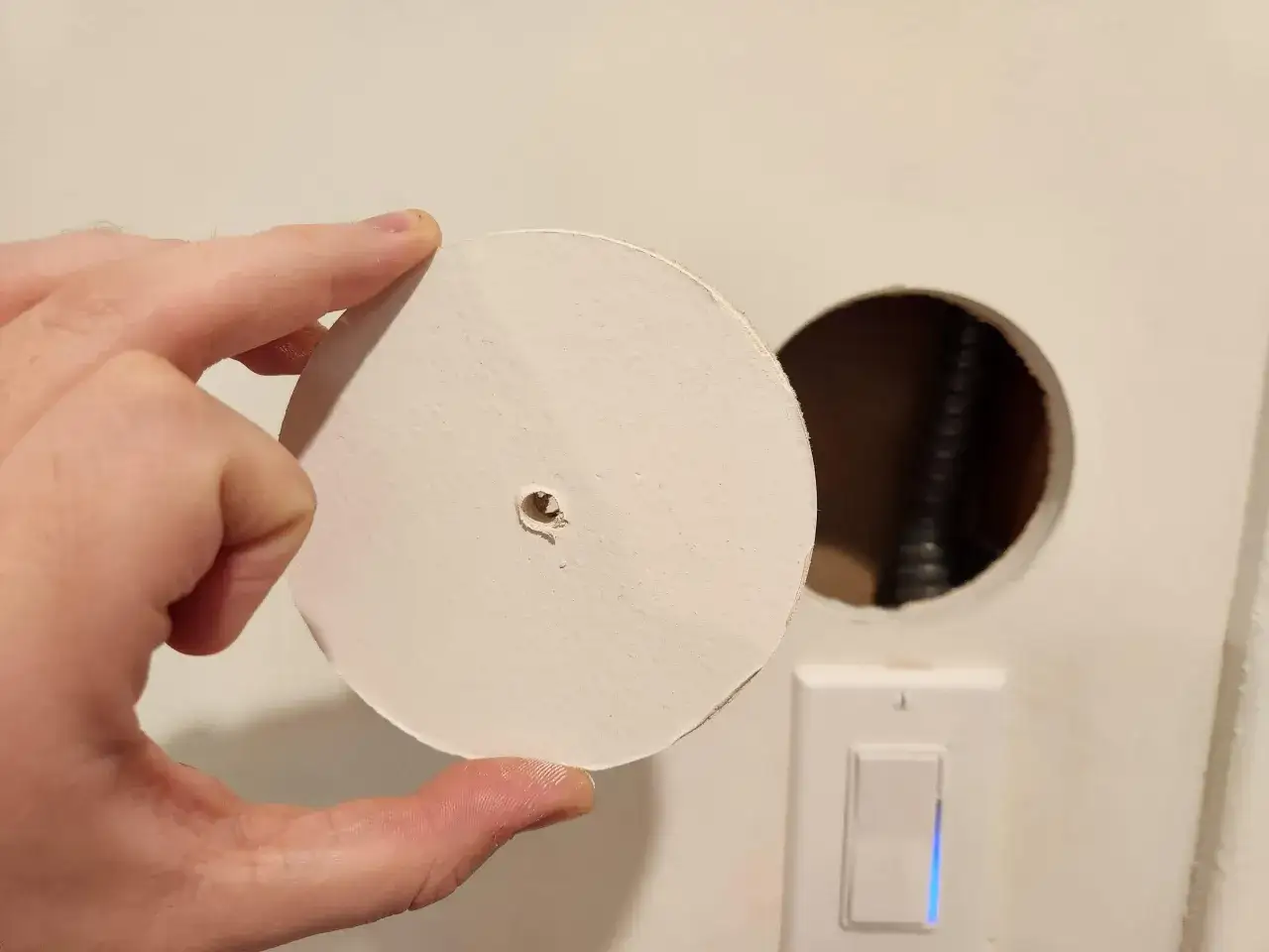Mano tiene un disco di cartongesso, pronto a riparare un buco nel cartongesso. Vicino, un interruttore con luce blu.