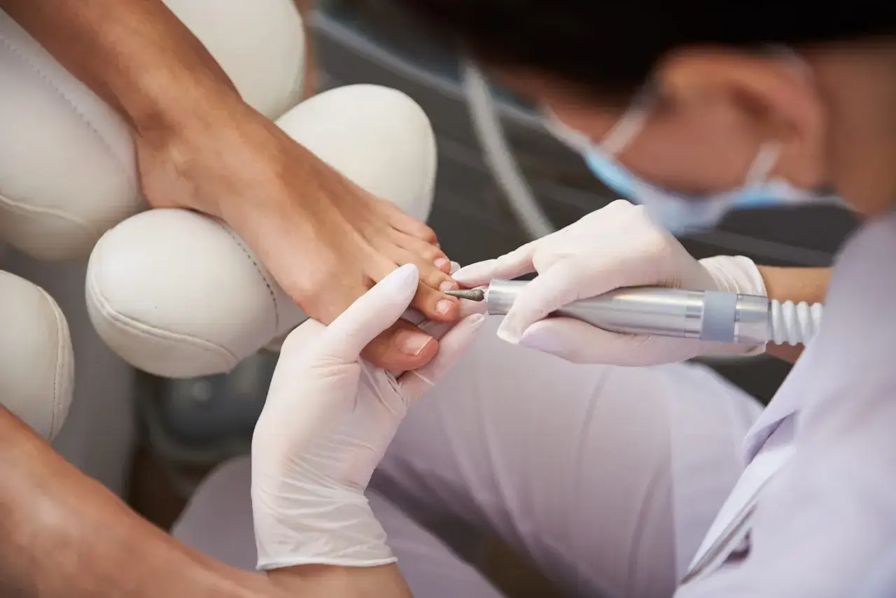 Profesjonalny pedicure frezarkowy w trakcie. Dłoń w rękawiczce delikatnie pracuje frezarką przy paznokciu stopy.