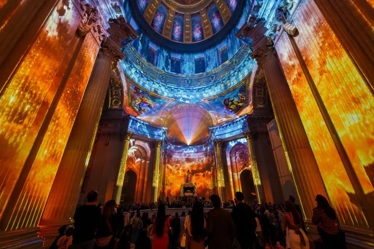 Spectacle lumineux projeté sur des colonnes dorées et une coupole ornée, créant une aura féerique. Une foule admire cette installation, oubliant les dates.
