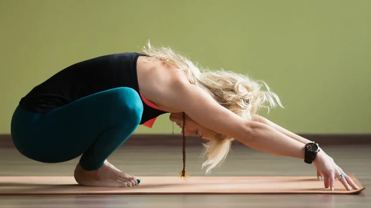 Kobieta w pozycji malasana na macie do jogi, z dłońmi wyciągniętymi do przodu, pogłębiając skłon.