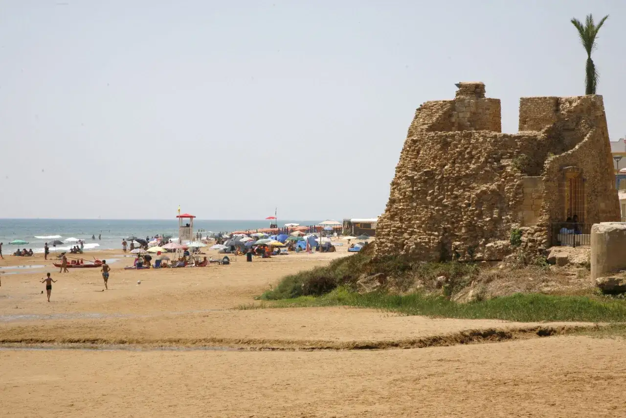 Spiaggia affollata con ombrelloni, bagnanti e un'antica torre di pietra. Vicino alla costa, si intravedono quelle che sembrano tre fontane naturali che sfociano nel mare.