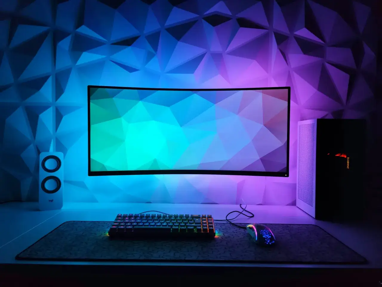 Un bureau gaming avec un écran incurvé, clavier RGB, souris et enceintes, le tout baigné de lumières bleues et violettes. Parfait comme fond ecran gaming.