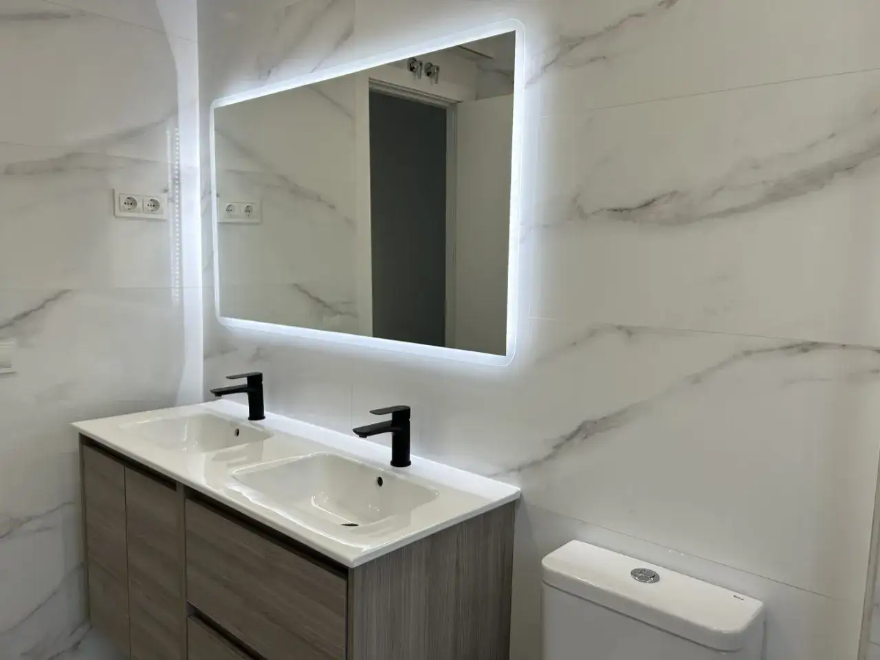 Moderno lavabo doble con grifería negra, espejo iluminado y revestimiento de mármol. Una solución ideal para reformar baño sin obras y darle un toque de estilo.