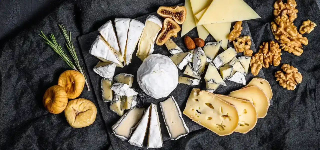 Un assortimento di formaggi, tra cui un cremoso formaggio morbido, fichi secchi, noci e rosmarino, su una tavola scura.