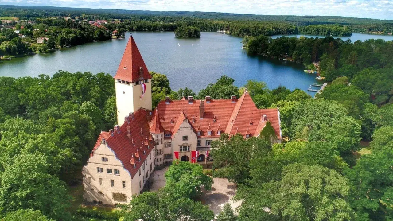 Zamek w Lubniewicach, otoczony zielenią, z czerwonymi dachami, g&oacute;ruje nad jeziorem.