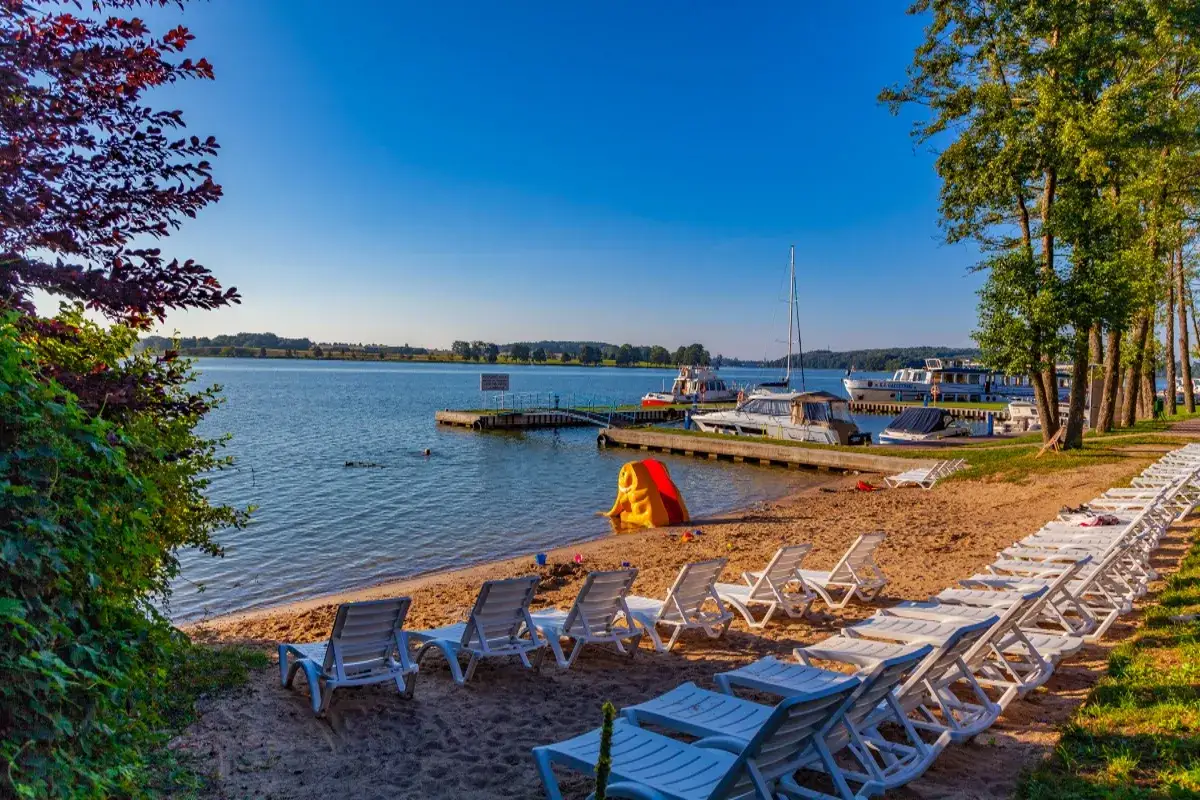 Piękny dzień nad jeziorem. Leżaki czekają na plaży, a łodzie cumują przy pomoście. Idealne miejsce na wypoczynek w mazurskich miejscowościach.
