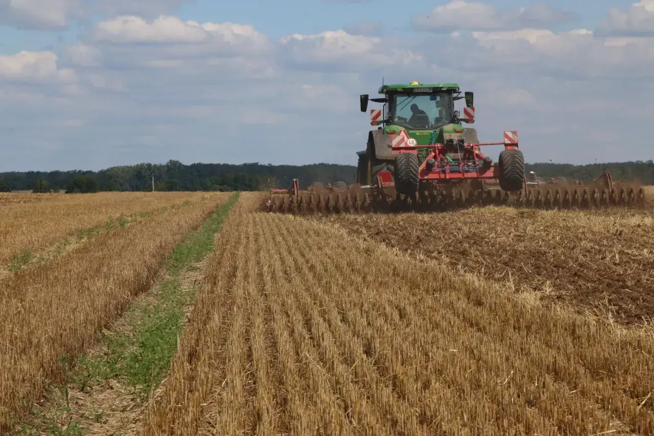 Zielony traktor z agregatem uprawowym, niczym buster, orze pole po żniwach. W tle las i błękitne niebo z chmurami.