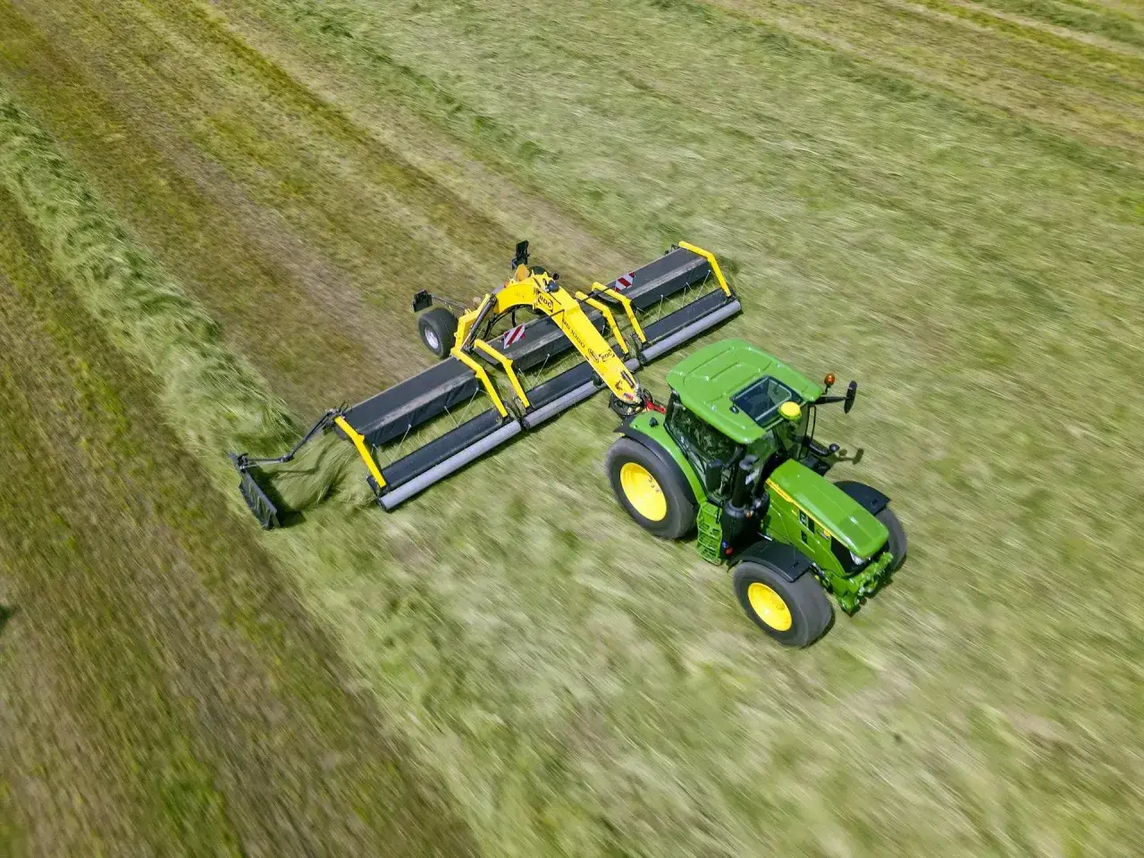 Zielony traktor z ż&oacute;łtą zgrabiarką taśmową pracuje na zielonym polu, zbierając skoszoną trawę.