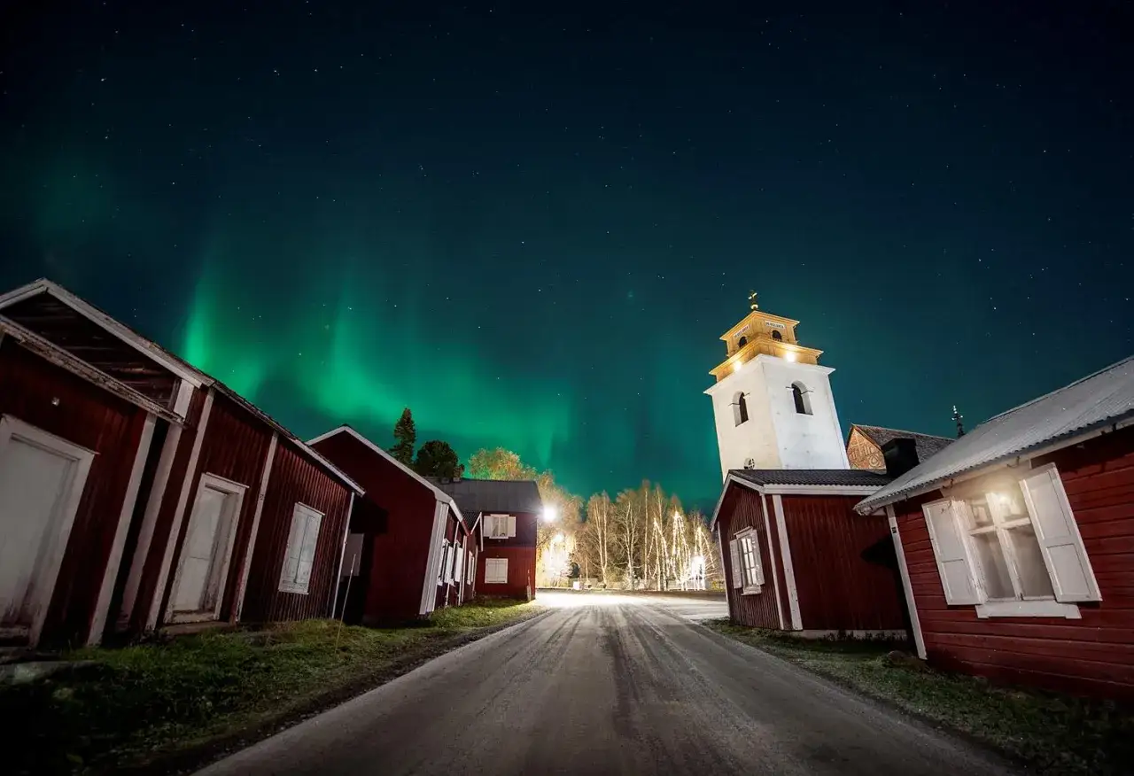 Kyrkstaden Luleå badar i norrskenets gröna sken. Röda hus och kyrktorn under en stjärnklar himmel.