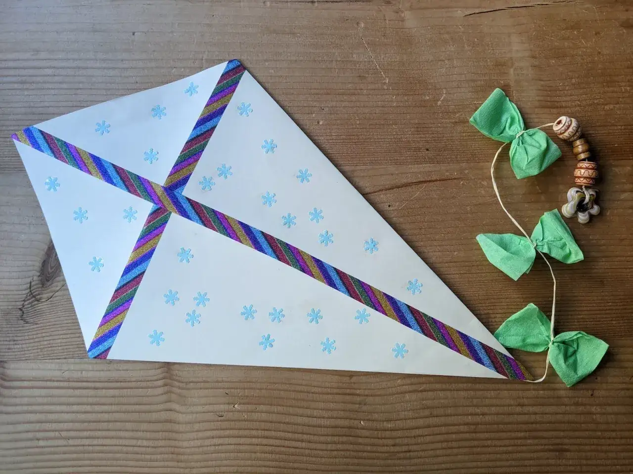 Bastelideen: Ein Drachen aus weißem Papier mit Schneeflocken und bunten Bändern, dazu eine Schnur mit grünen Schleifen und Holzperlen.
