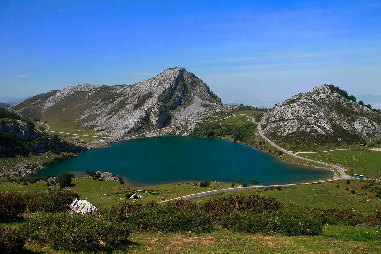Lago azul rodeado de montañas rocosas y verdes prados. Un camino serpentea junto al agua, ideal para descubrir qué hacer en Cangas de Onís.