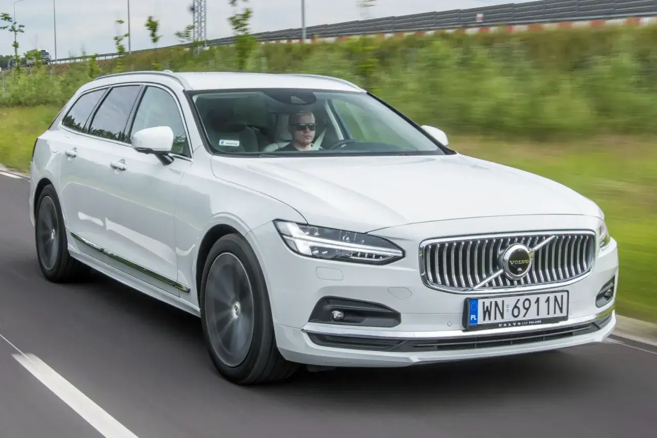 Białe Volvo V90 Cross Country w ruchu drogowym. Kierowca w okularach przeciwsłonecznych. Wiele osób szuka volvo v90 cross country opinie.