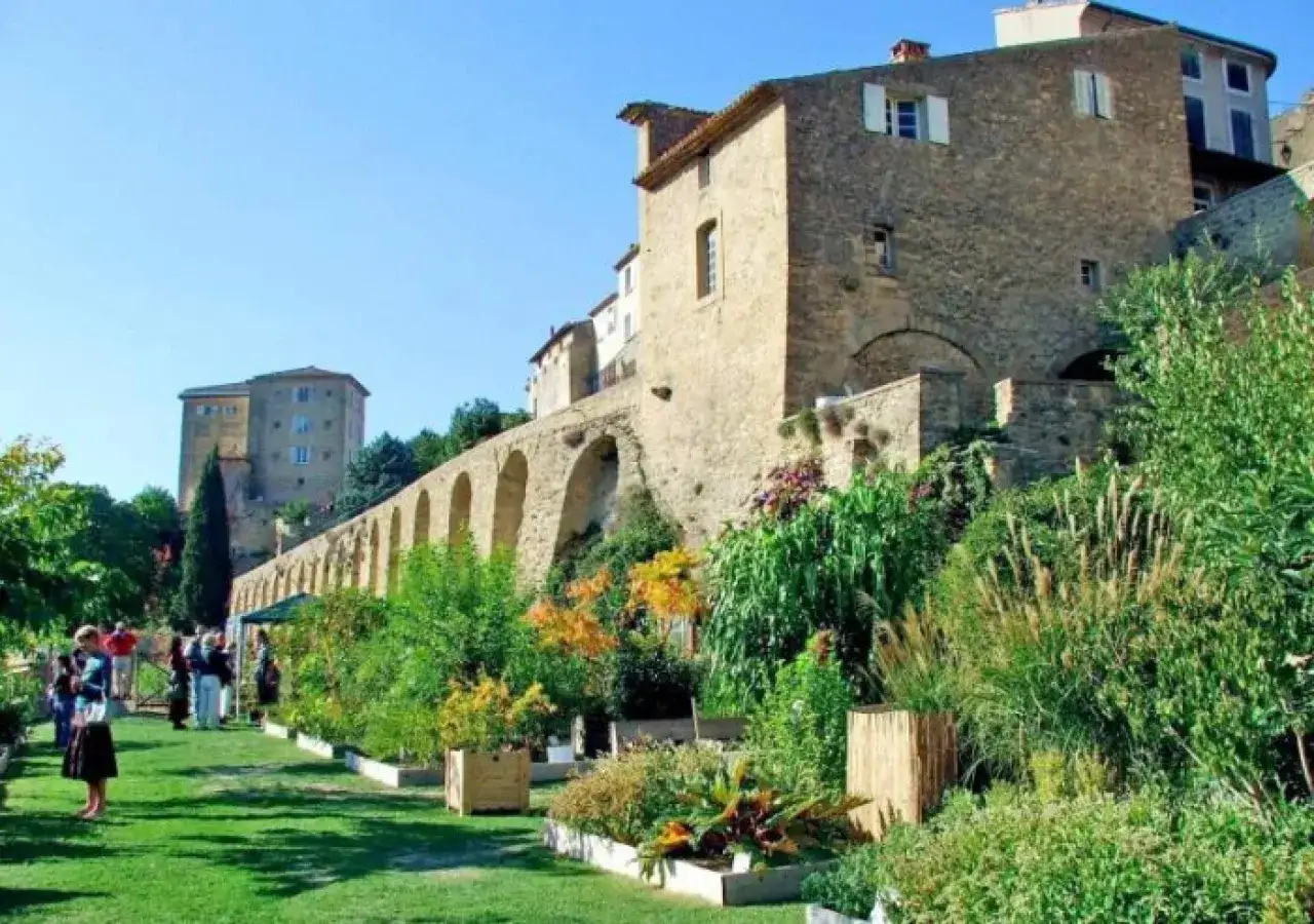 Jardin fleuri devant le **ch&acirc;teau de Lauris**, avec ses remparts vo&ucirc;t&eacute;s et ses b&acirc;timents en pierre sous un ciel bleu.