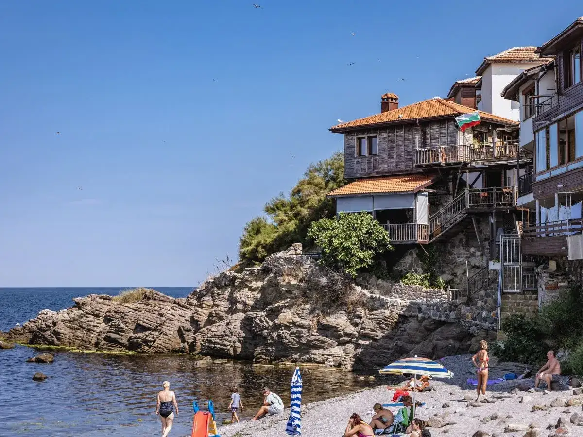 Słoneczny dzień na plaży w bułgarskim miasteczku, przypominającym urokliwe zakątki **Marseille**. Ludzie odpoczywają, a domy w stylu śródziemnomorskim wznoszą się na klifie.