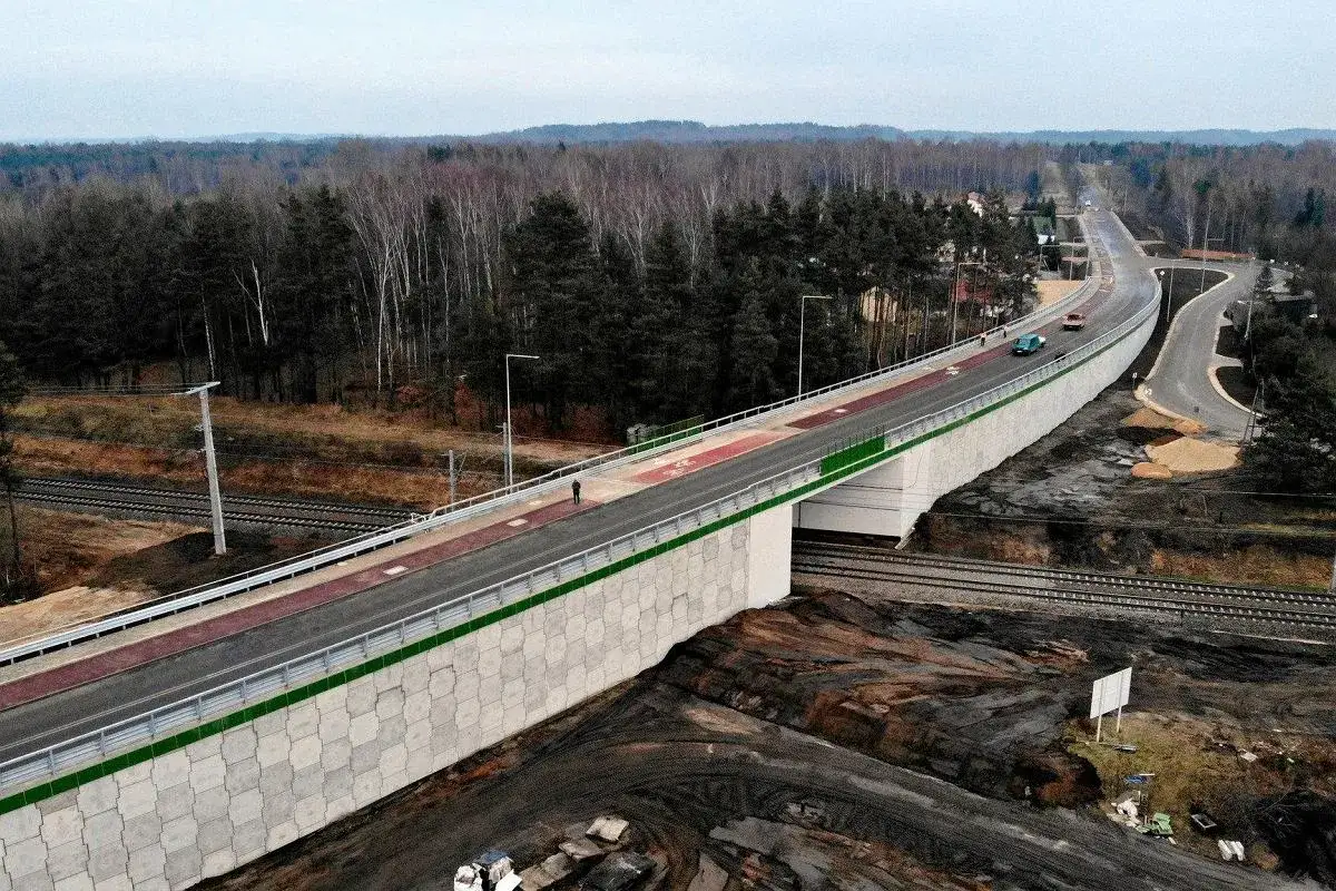 Nowy most nad torami kolejowymi, z drogą i ścieżką rowerową, otoczony lasem.
