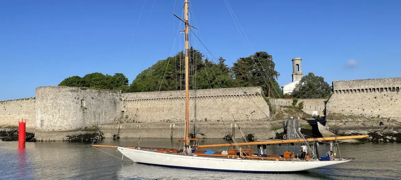 Un voilier blanc, le Moonbeam 4, navigue près d'une ancienne forteresse sous un ciel bleu.