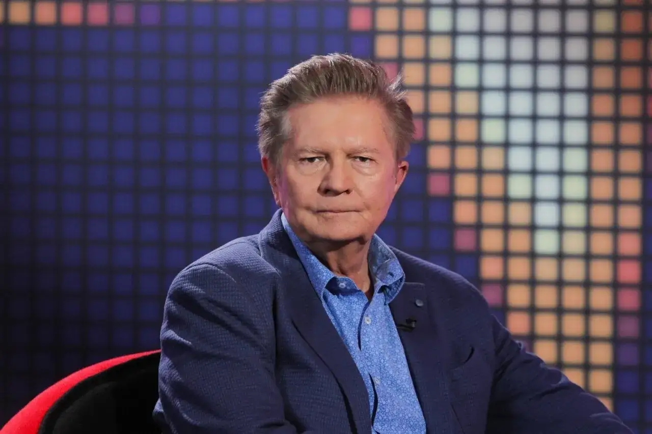 Krzysztof Szewczyk w niebieskiej marynarce i koszuli, siedzi na tle kolorowej, pikselowej ściany.