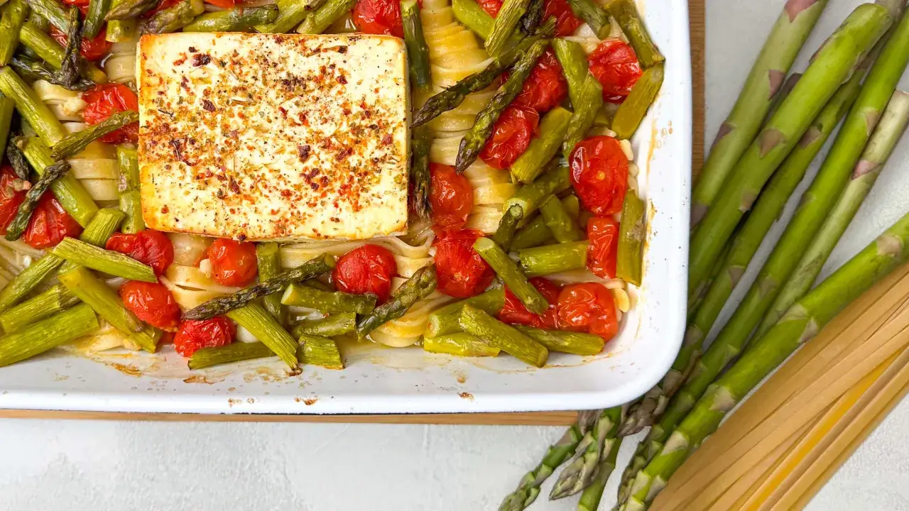 Eine Auflaufform mit Spargel, Kirschtomaten und Pasta, gekrönt von einem Block Feta. Daneben liegen frischer Spargel und ungekochte Pasta.