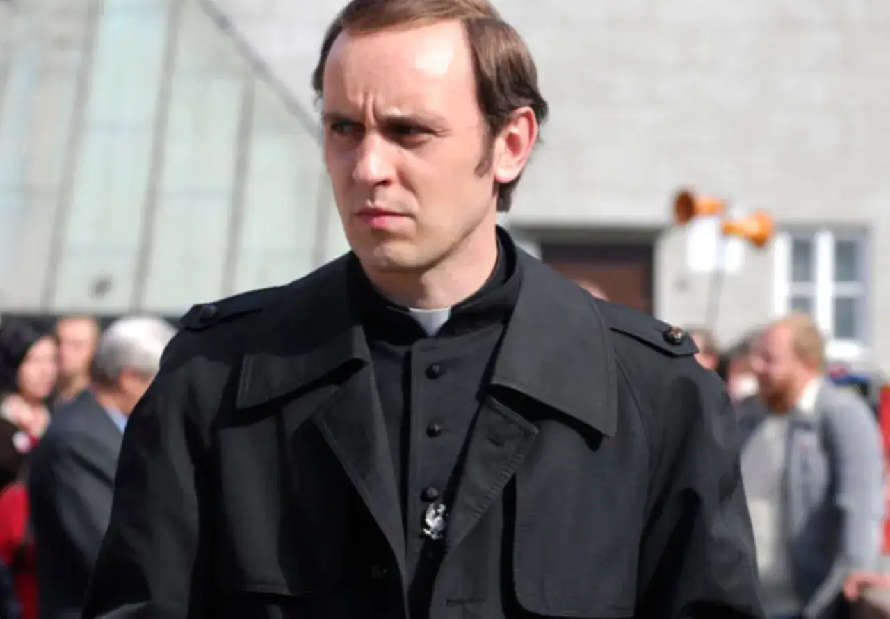 Aktor w czarnym płaszczu i sutannie, który grał Popiełuszkę, patrzy w bok z powagą.