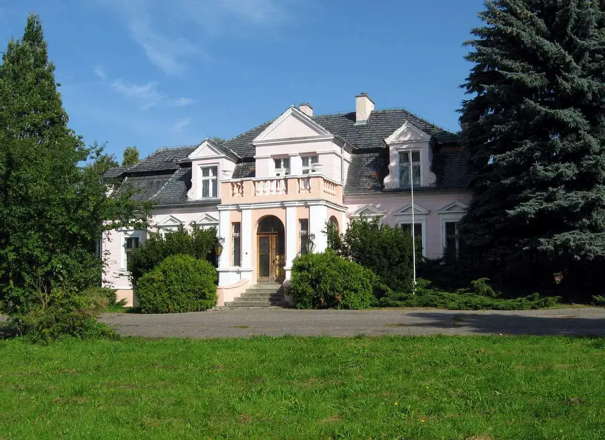 R&oacute;żowy dworek Wybickich z balkonem nad wejściem, otoczony zielenią i dużym drzewem iglastym.