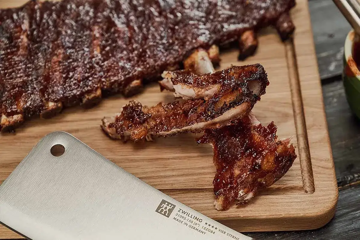 Fertig marinierte Spareribs werden auf einem Holzbrett pr&auml;sentiert, bereit zum Genuss nach dem Grillen auf dem Gasgrill. Ein Messer liegt daneben.
