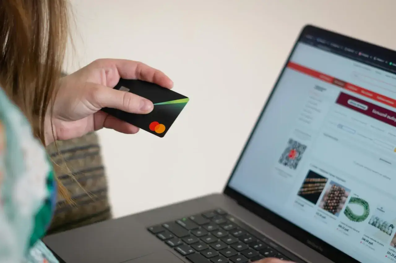 Karta płatnicza w dłoni, ekran laptopa z ofertami. Chcesz wiedzieć, jak sprawdzić do kogo należy numer konta bankowego?