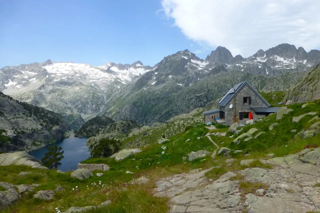 Refugi com de rubio, un refugio de piedra junto a un lago azul, rodeado de monta&ntilde;as nevadas y rocas cubiertas de hierba.