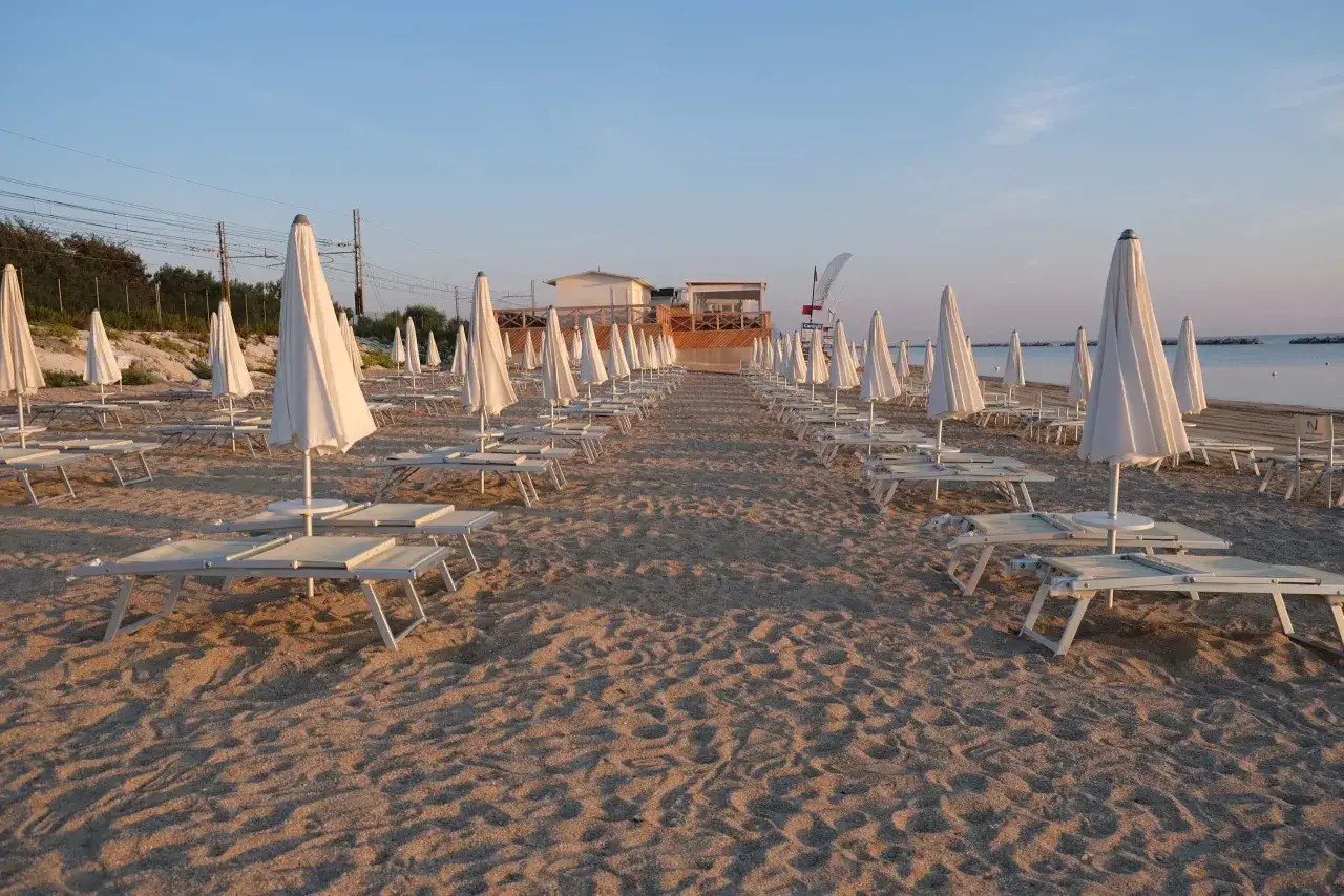 File di lettini e ombrelloni chiusi su una spiaggia dorata, pronti per accogliere i bagnanti in un **mare bello** nelle **Marche**.