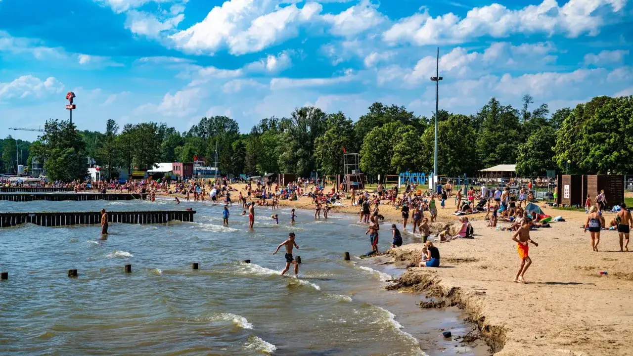 Tłum ludzi cieszy się słońcem i kąpielą na piaszczystej plaży. Woda jest spokojna, a drzewa tworzą zielone tło. To idealne miejsce na relaks nad najcieplejszym jeziorem w Polsce.