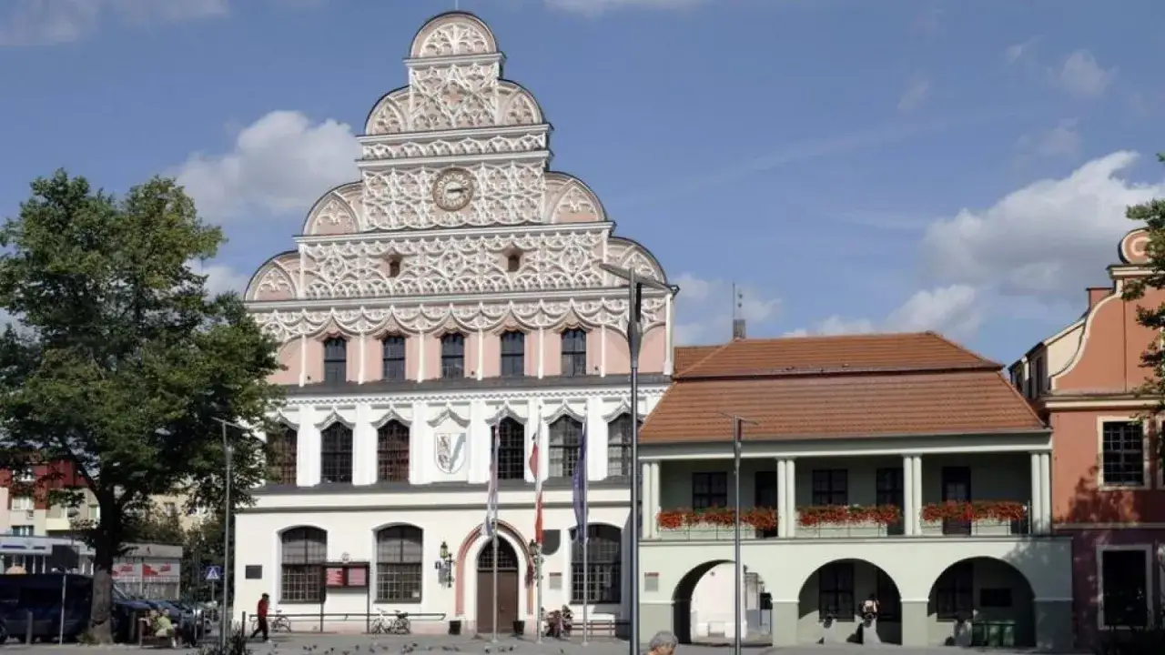 Ratusz w mieście nad Iną, z bogato zdobioną fasadą i zegarem, góruje nad placem. Obok zielony budynek z arkadami.
