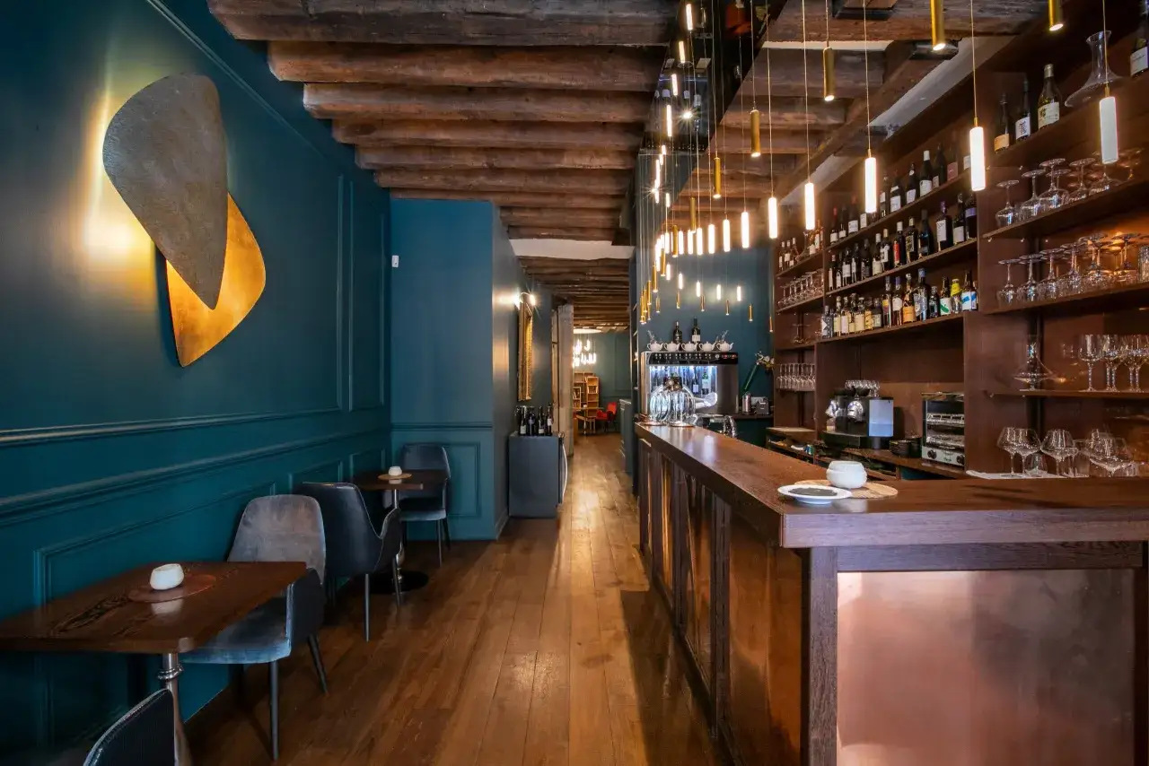 Ambiance chaleureuse d'un bar à vin parisien avec ses poutres apparentes, son mur bleu canard et son comptoir en bois.