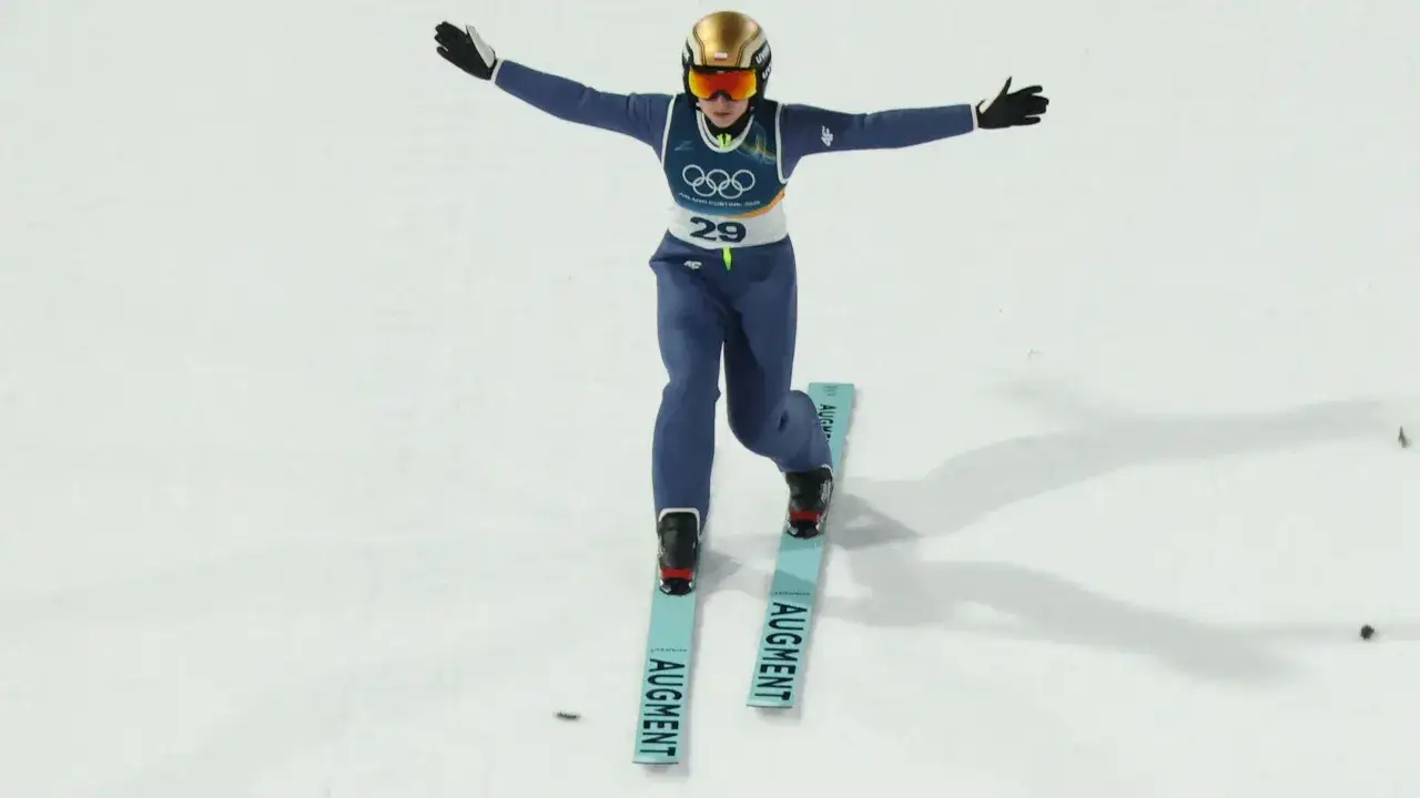 Skoczek narciarski w złoto-niebieskim kombinezonie i kasku, z rękami rozłożonymi na boki, gotowy do lotu. Widoczne logo igrzysk olimpijskich Milano Cortina 2026.