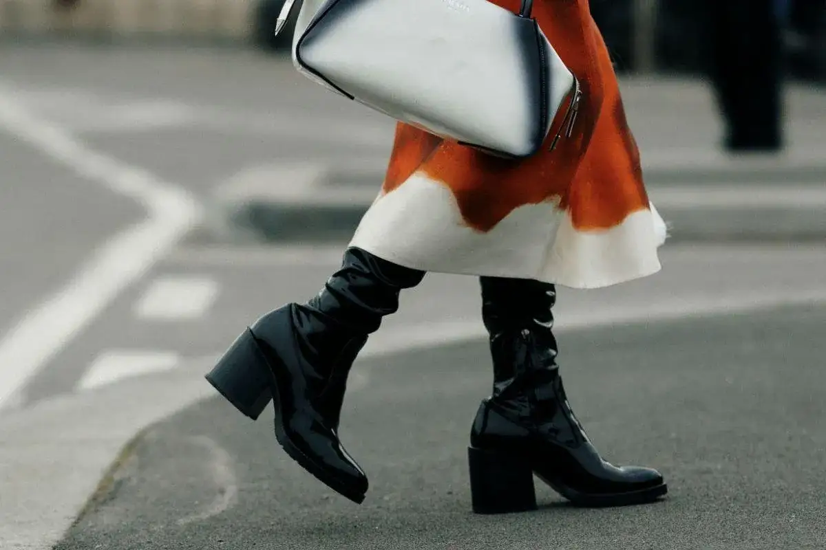 Mujer con falda naranja y blanca y unas **botas** negras brillantes con tac&oacute;n grueso, caminando por la calle.