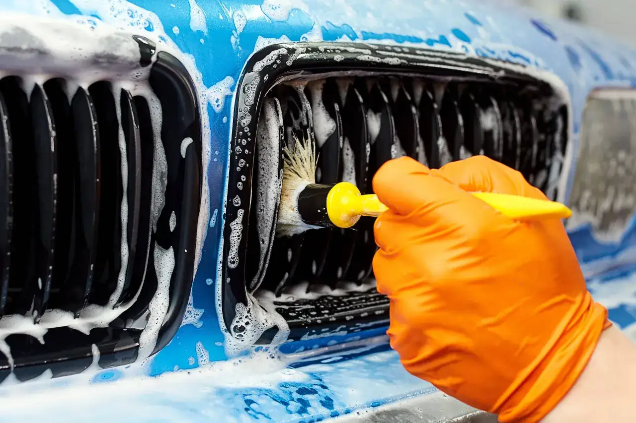 Mano con guante naranja usa cepillo para limpiar la parrilla de un coche azul cubierto de espuma. Detalle de c&oacute;mo limpiar la carrocer&iacute;a del coche.