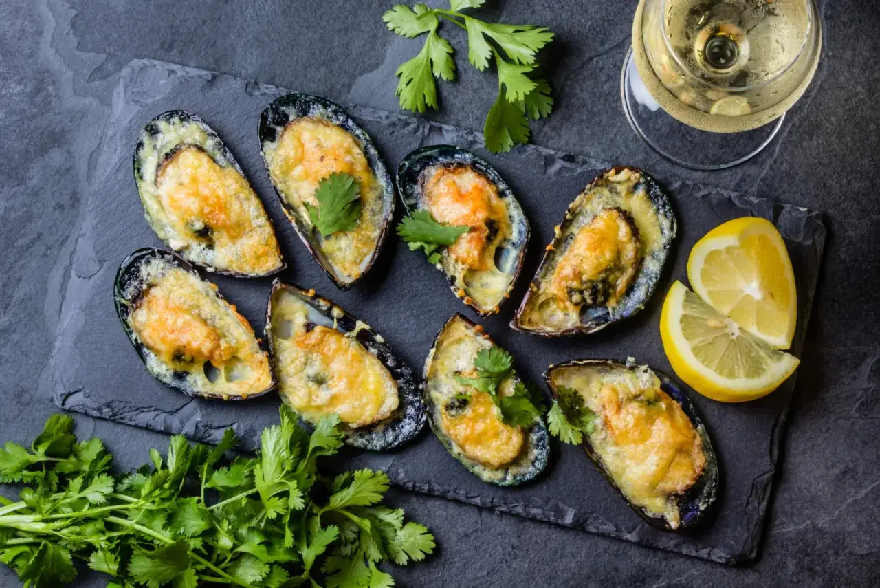Huit moules au barbecue gratin&eacute;es au fromage, garnies de coriandre fra&icirc;che, servies avec des quartiers de citron et un verre de vin blanc.