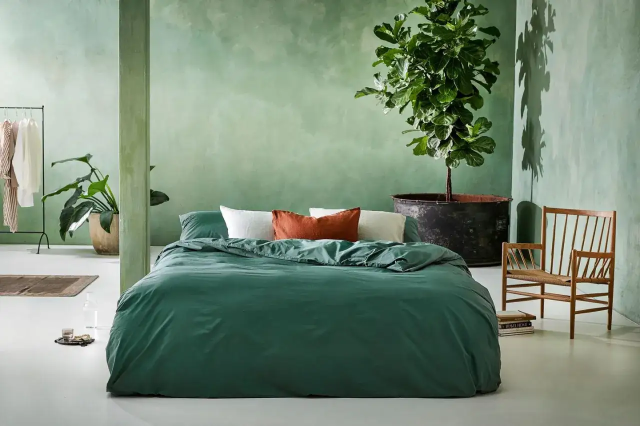 Grünes Schlafzimmer mit erdigen Akzenten. Die Schlafzimmer Farbgestaltung wirkt beruhigend und natürlich, mit einer großen Zimmerpflanze und einem Holzsessel.