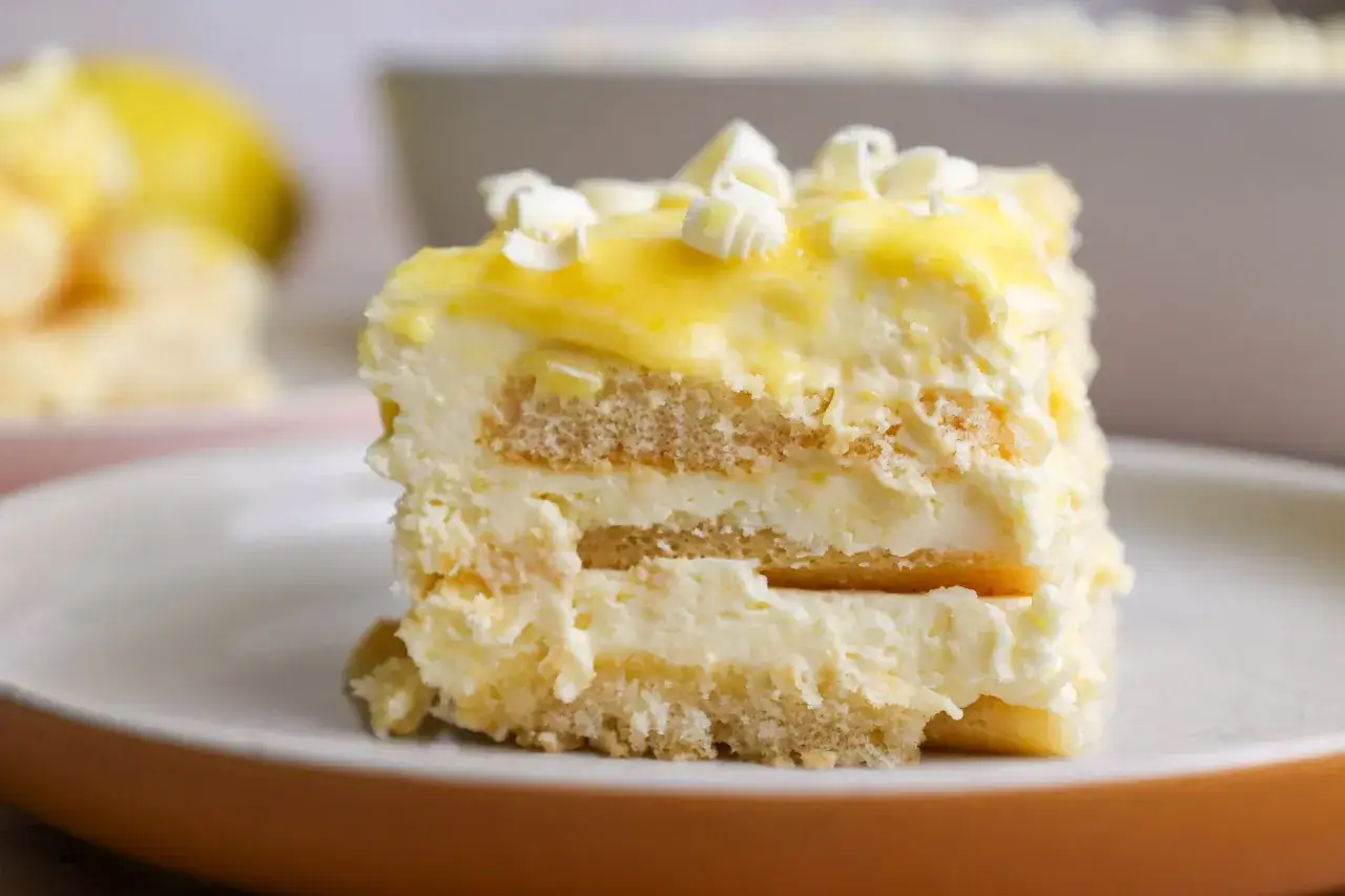 Rebanada de tiramisú de limón, con capas de bizcocho y crema, cubierta de glaseado amarillo y virutas de chocolate blanco.